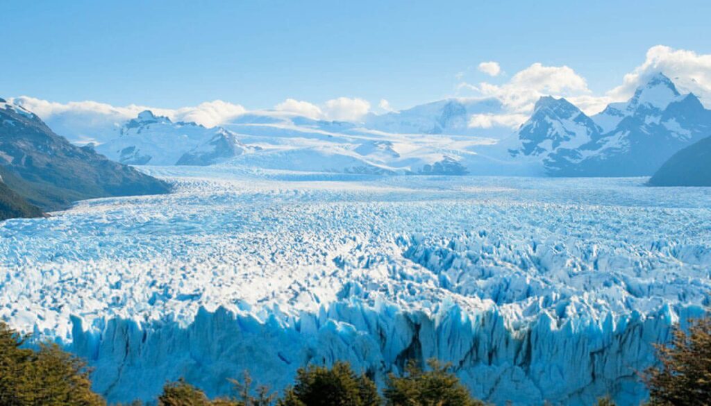 Things to do in El Calafate Patagonia 3 A vast glacier stretches across the Patagonian landscape near El Calafate, framed by jagged snow-capped mountains and scattered green trees under a bright blue sky with some clouds.