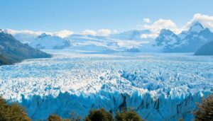 A vast glacier stretches across the Patagonian landscape near El Calafate, framed by jagged snow-capped mountains and scattered green trees under a bright blue sky with some clouds.