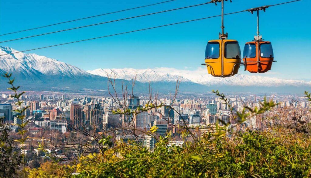 Two colorful cable cars glide above a city of modern buildings in South America, framed by snow-capped mountains, clear blue sky, and lush green foliage—one of the Top 5 scenic urban cable car views.