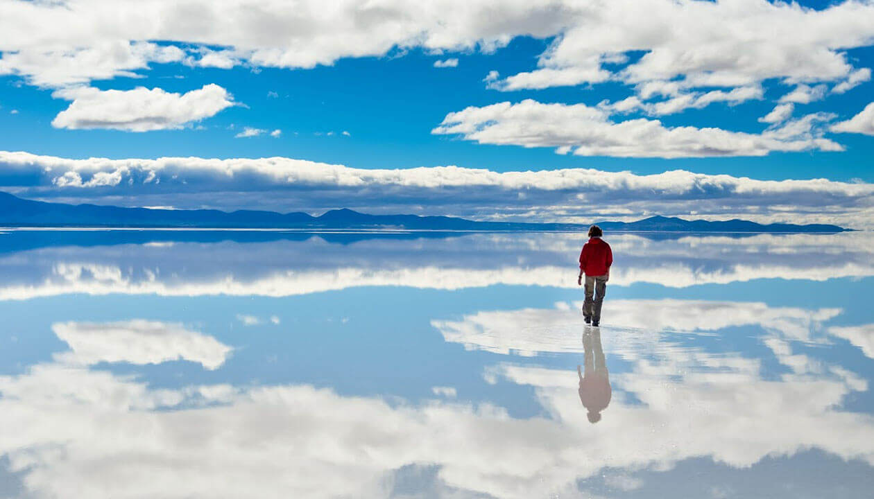 A person in a red jacket walks alone on a mirror-like salt flat beneath a blue sky with scattered clouds and distant mountains—one of Bolivia’s top 5 places to visit.