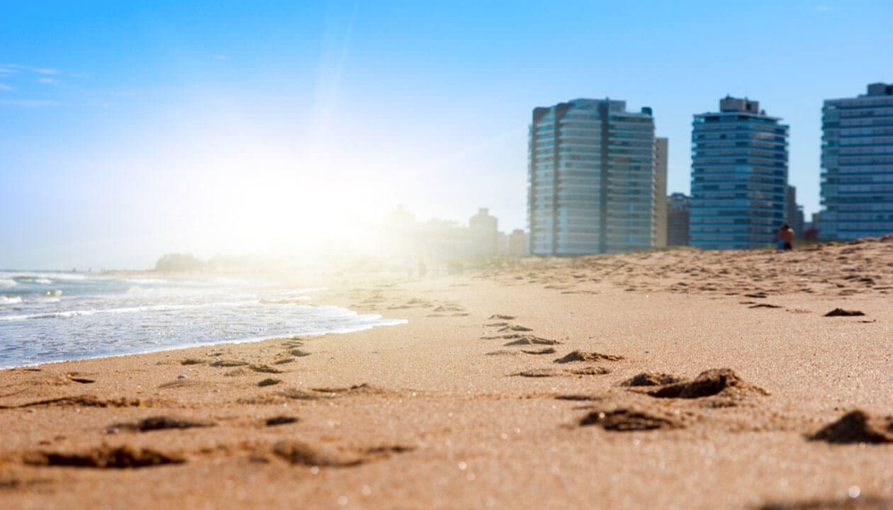Footprints in the sand lead toward the ocean on a sunny beach in Montevideo, with a few modern high-rise buildings in the background under a clear blue sky—perfect for travel and discovering new things to do.