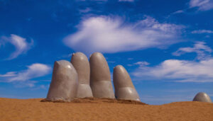 A large sculpture of five giant fingers emerges from sandy ground under a bright blue sky with scattered clouds, making it one of Punta del Este’s top attractions and a must-see when searching for things to do in the area.