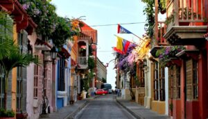 A narrow street in Cartagena lined with colorful colonial buildings, balconies decorated with flowers, and Colombian flags hanging under a clear sky—discovering sights like this is one of the best things to do in Colombia. A red car is parked in the distance.