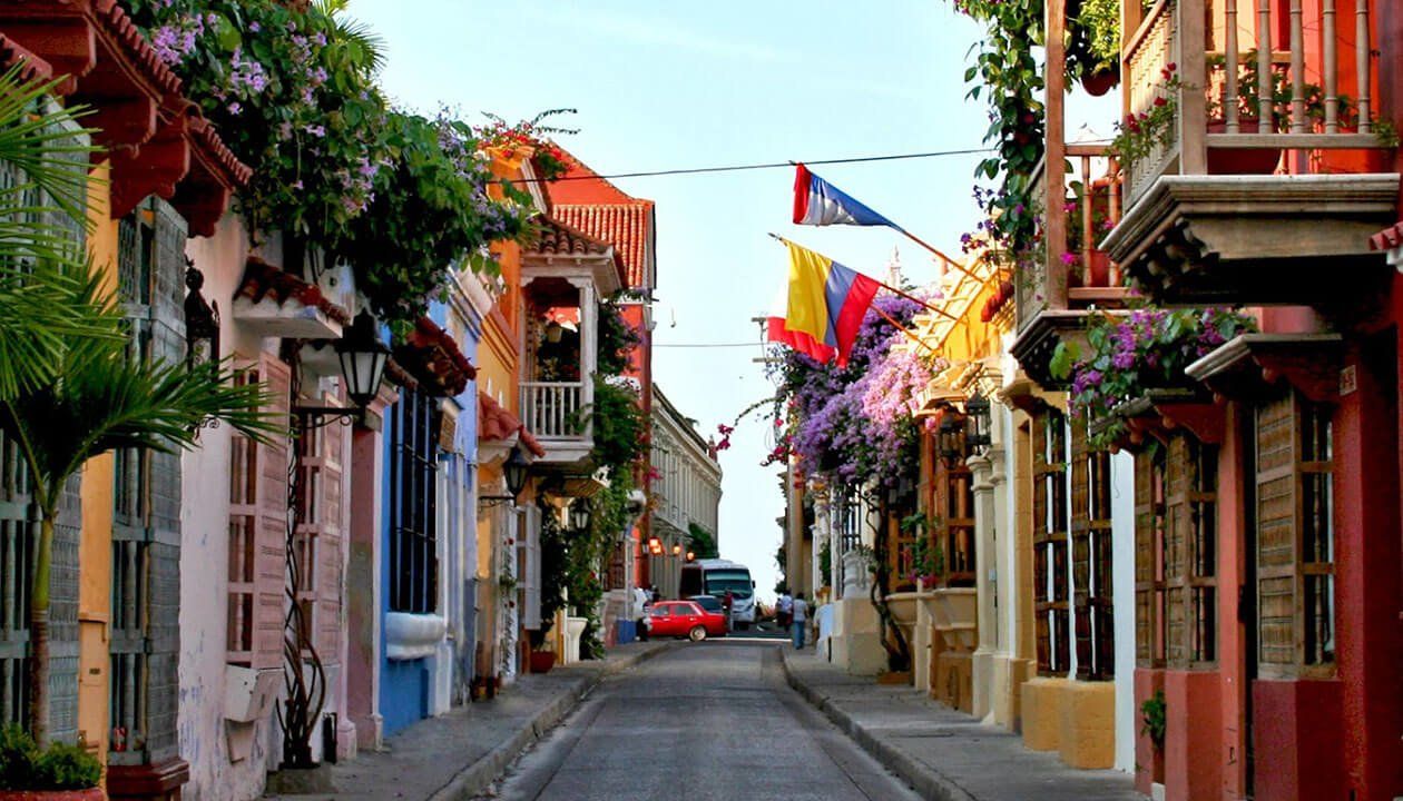 A narrow street in Cartagena lined with colorful colonial buildings, balconies decorated with flowers, and Colombian flags hanging under a clear sky—discovering sights like this is one of the best things to do in Colombia. A red car is parked in the distance.