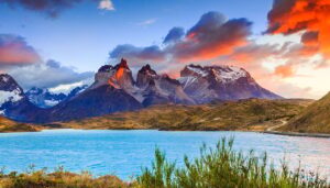 A turquoise lake in Chile sits before rugged, snow-capped mountains under a dramatic sky with orange and pink clouds, surrounded by lush green vegetation—a breathtaking travel destination and one of the must-see places to visit.