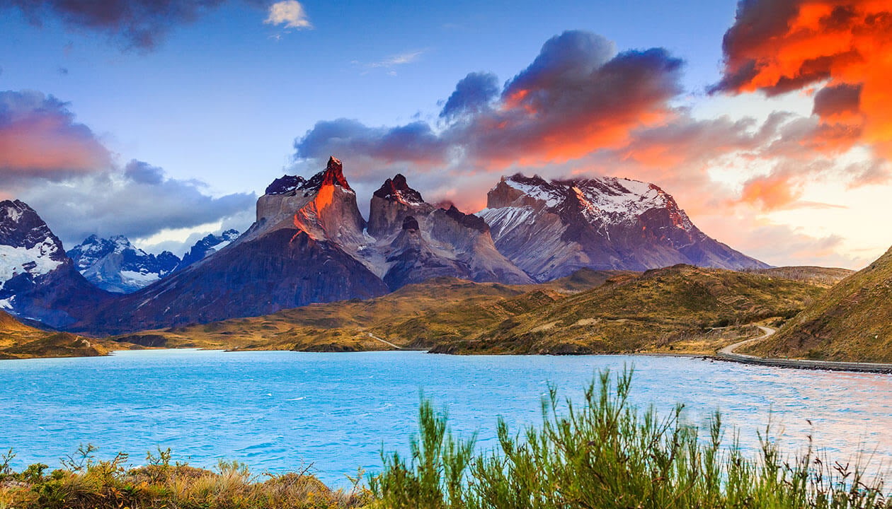 A turquoise lake in Chile sits before rugged, snow-capped mountains under a dramatic sky with orange and pink clouds, surrounded by lush green vegetation—a breathtaking travel destination and one of the must-see places to visit.