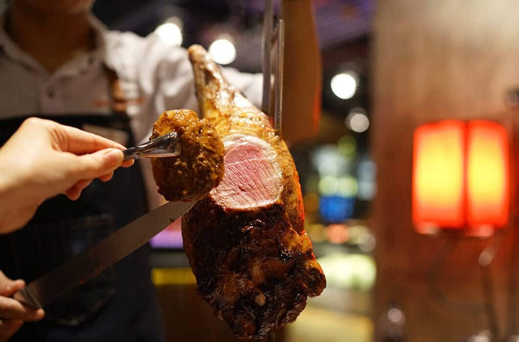 A server in a white shirt carves roast meat tableside, highlighting classic Brazil attractions. Another hand uses tongs to hold the sliced portion, set against a backdrop of warm, blurred lights and a glowing red lamp—authentic Brazil travel at its best.