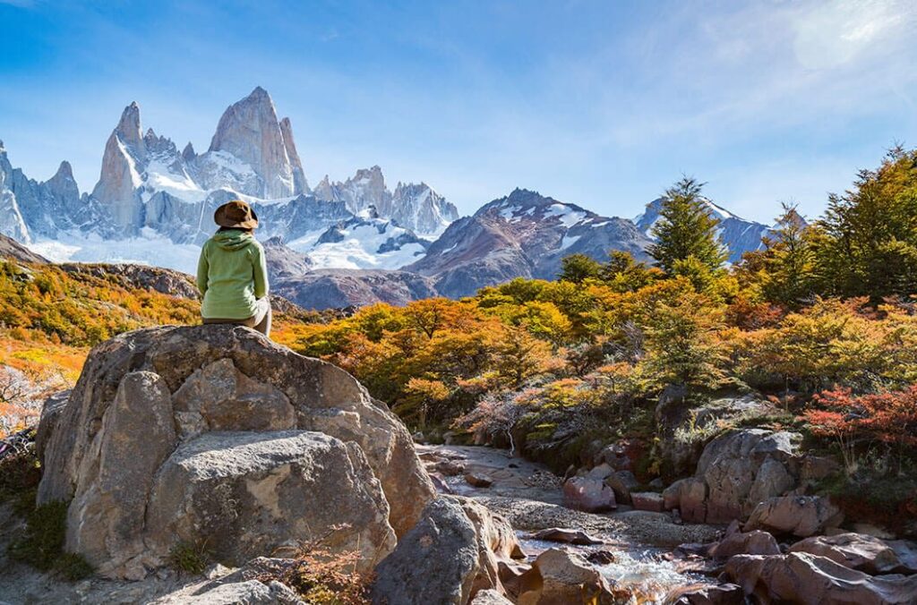 A person in a hat and green jacket sits on a large rock in Patagonia, gazing at snow-capped mountains. Colorful autumn trees and a clear blue sky complete this stunning travel scene.