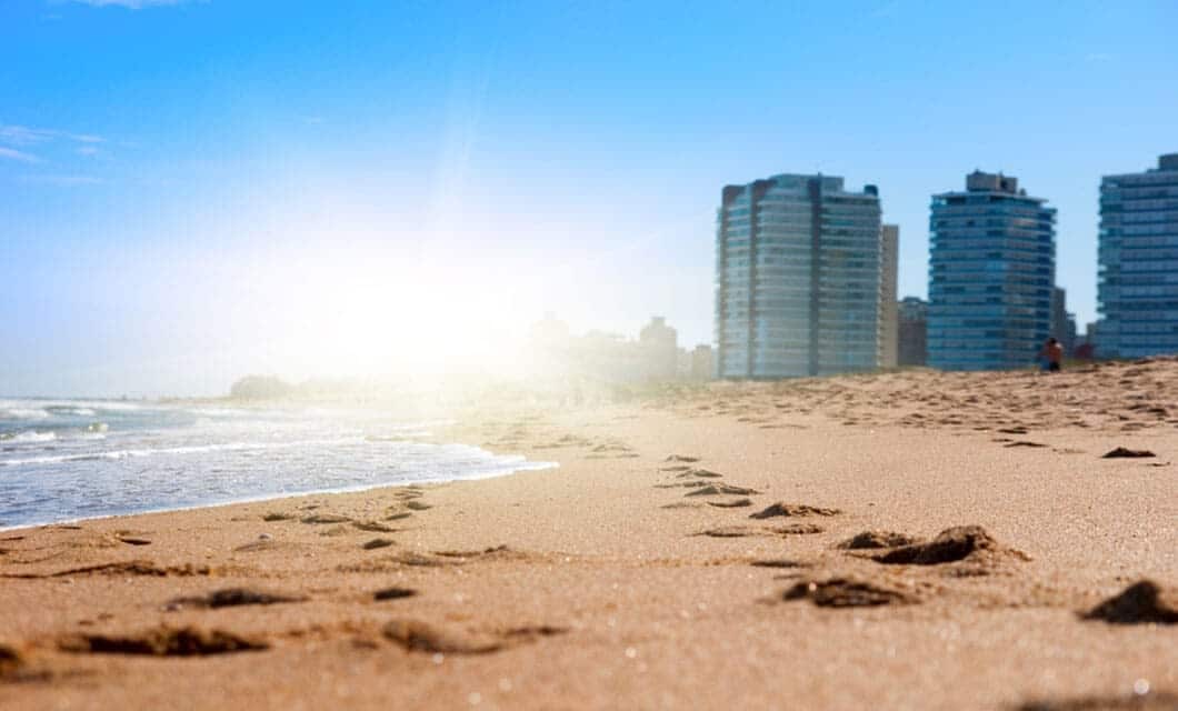 Footprints in the sand lead toward the shoreline on a sunny beach, with gentle waves and tall modern buildings in the background—a perfect scene for those seeking unforgettable Uruguay travel experiences.
