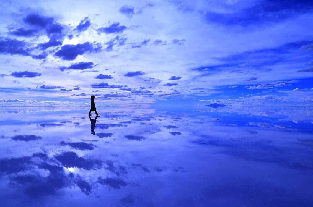 A person walks alone on a vast, reflective surface under a blue sky filled with clouds, creating a calm and surreal scene—one of the iconic Bolivia attractions for unforgettable Bolivia travel experiences.