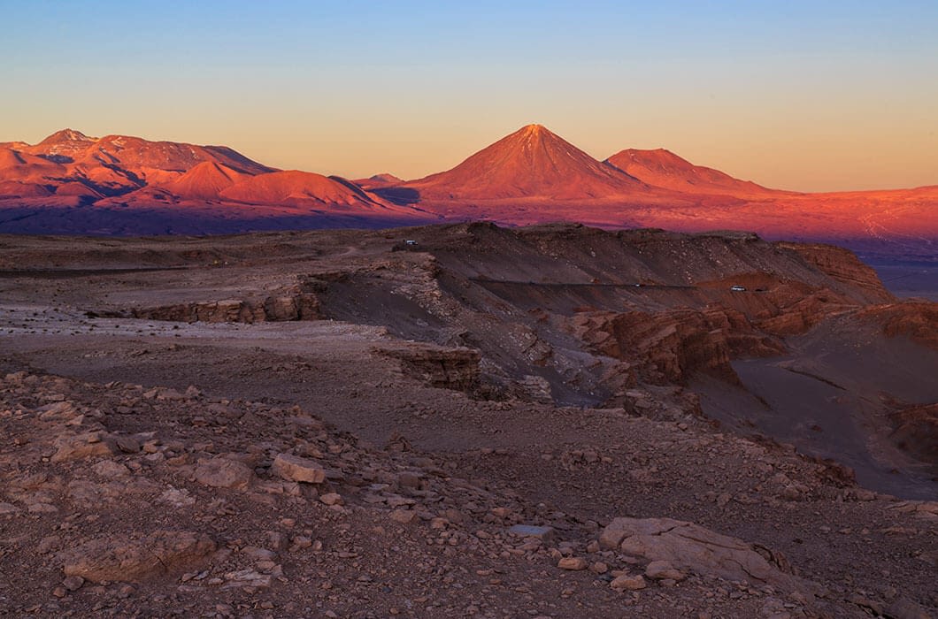 A rocky desert landscape with distant mountains and a prominent volcano, all bathed in warm sunset light under a clear sky—one of the stunning Bolivia attractions for any Bolivia travel adventure.