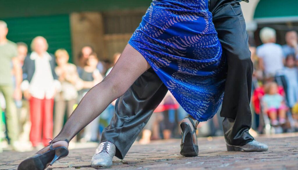 A couple dances the tango outdoors—a scene Argentina is famous for. The woman in a blue dress and fishnet stockings, the man in dark trousers and silver shoes, their legs entwined dramatically against a blurred South America travel crowd.