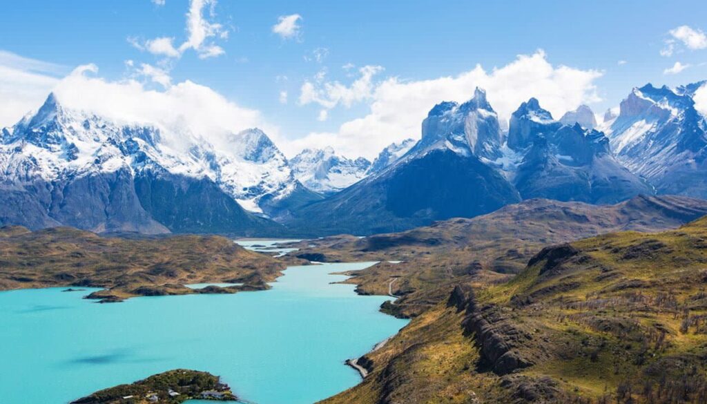 Snow-capped mountains tower over a bright turquoise lake, with rolling green hills and rugged terrain in the foreground—an unforgettable view among the best places in Patagonia, perfect for unforgettable Patagonia travel adventures.