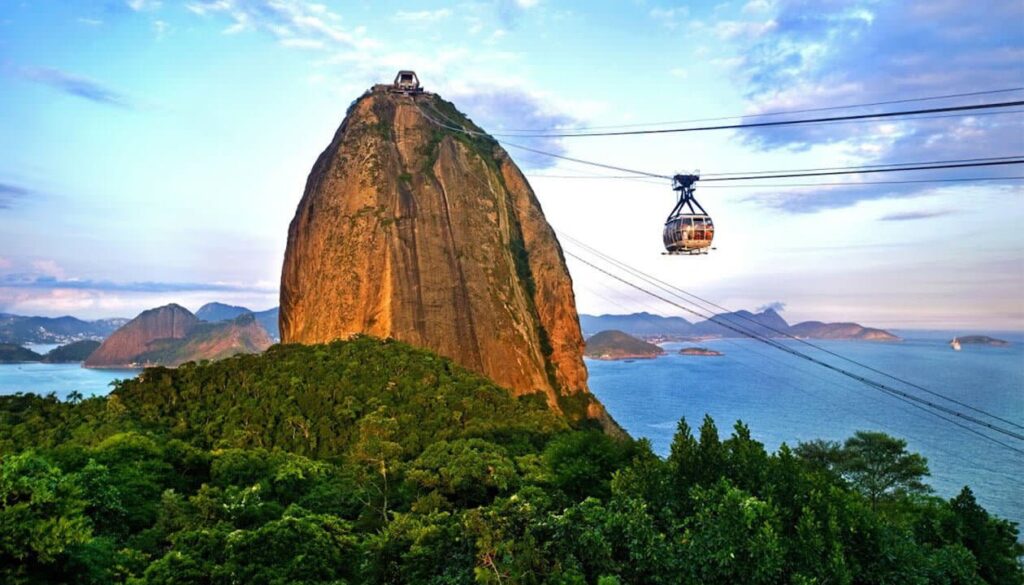 A cable car ascends toward Sugarloaf Mountain, a tall, rounded rock formation surrounded by lush greenery, with blue sky and ocean in the background—one of the most iconic Brazil attractions representing Brazil culture in Rio de Janeiro.