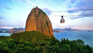 A cable car ascends toward Sugarloaf Mountain, a tall, rounded rock formation surrounded by lush greenery, with blue sky and ocean in the background—one of the most iconic Brazil attractions representing Brazil culture in Rio de Janeiro.