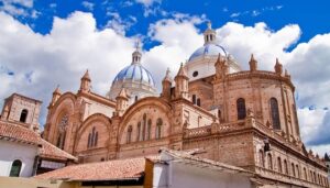 Large historic cathedral in Ecuador with tan stone walls and three striking blue-and-white domes under a bright blue sky with fluffy clouds, seen from a low angle. Rooftops of nearby buildings appear in the foreground.