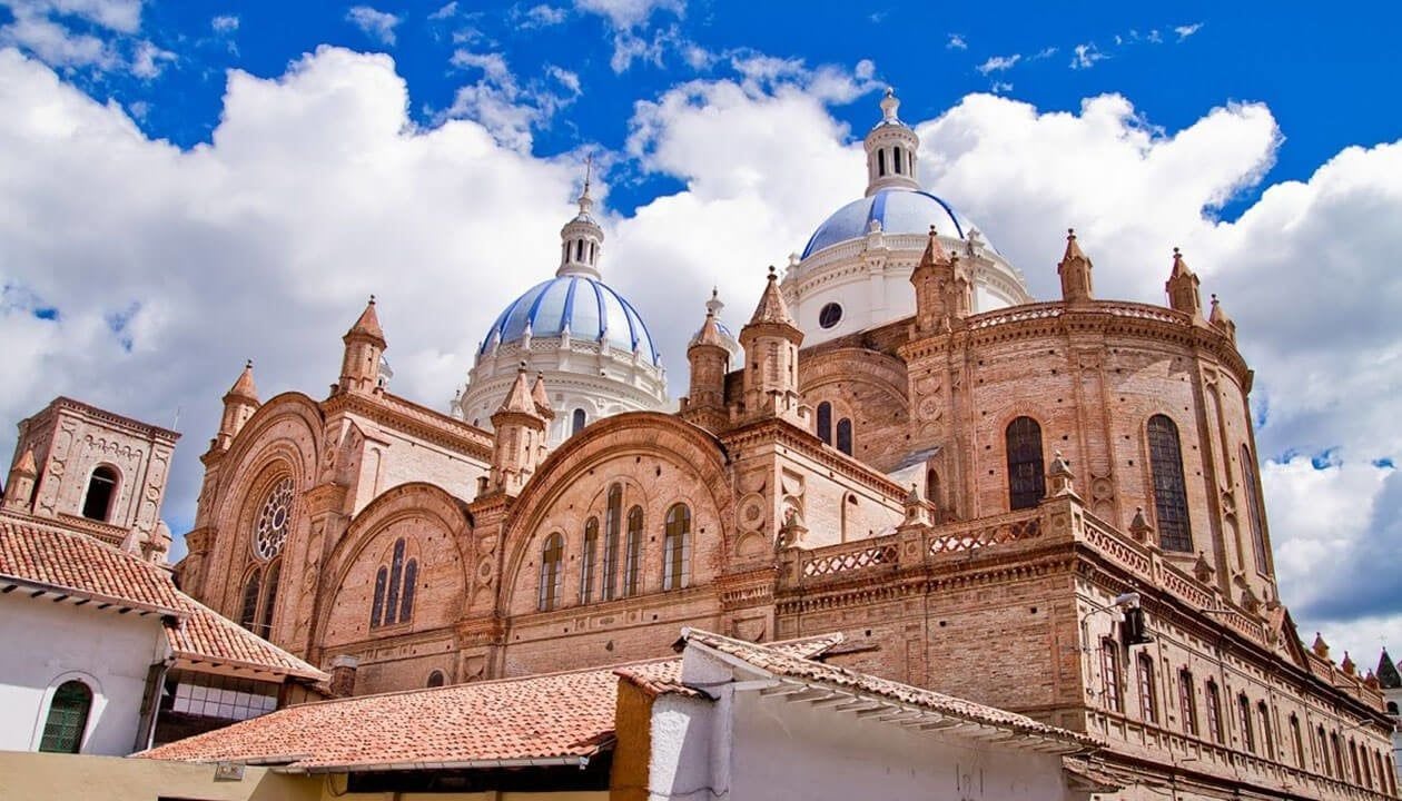 Large historic cathedral in Ecuador with tan stone walls and three striking blue-and-white domes under a bright blue sky with fluffy clouds, seen from a low angle. Rooftops of nearby buildings appear in the foreground.