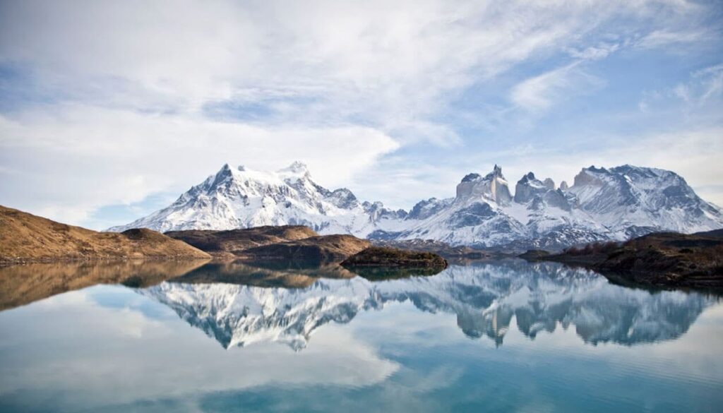 What is Patagonia Famous for? 6 Famous snow-capped mountains reflected in a calm, clear lake under a partly cloudy sky, with brown grassy hills in the foreground—this breathtaking view captures the essence of Patagonia.