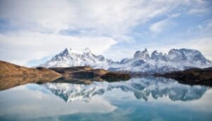 Famous snow-capped mountains reflected in a calm, clear lake under a partly cloudy sky, with brown grassy hills in the foreground—this breathtaking view captures the essence of Patagonia.