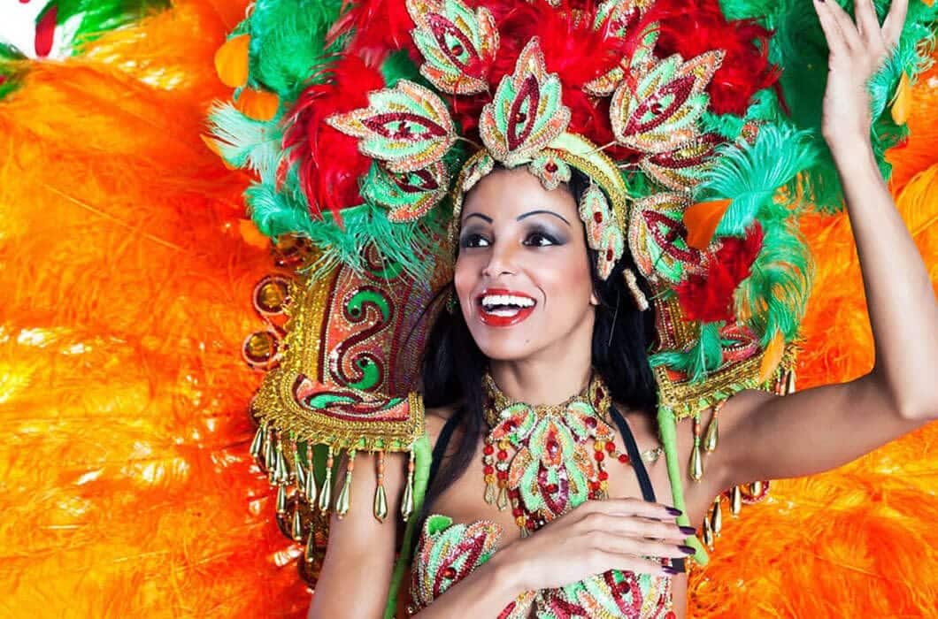 A smiling woman in a colorful, elaborate carnival costume with vibrant feathers in green, red, and orange. She wears a decorated headdress and jewelry, raising one arm in celebration—capturing the spirit of Brazil attractions.
