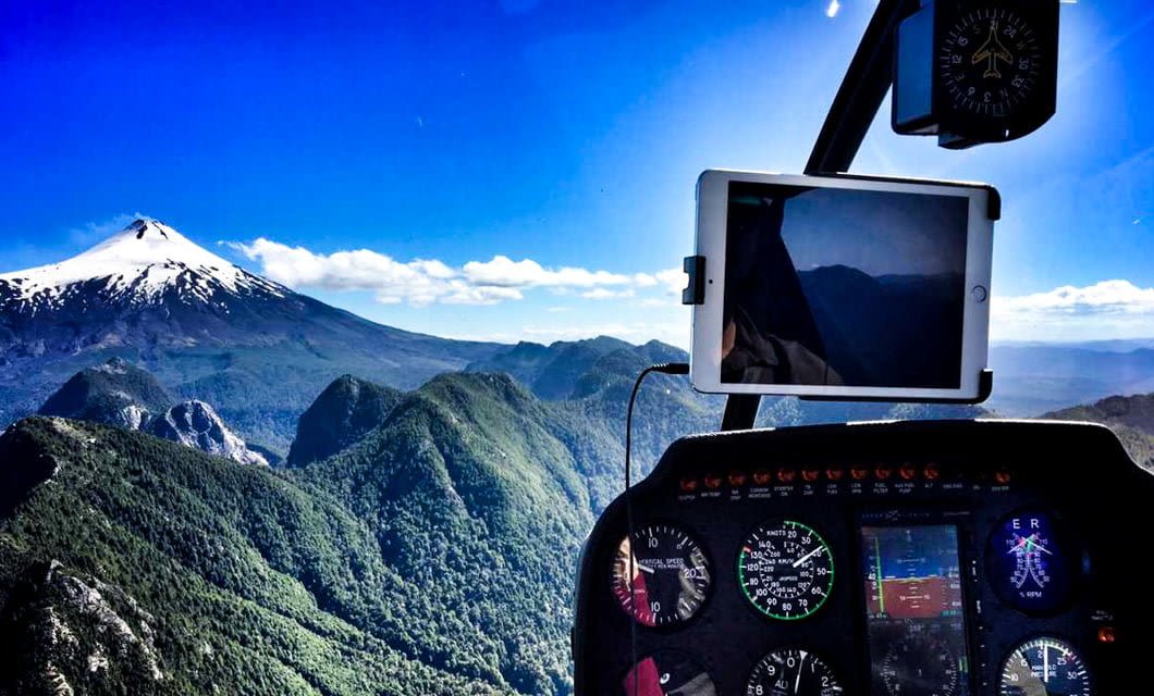 View from inside a helicopter cockpit flying over green mountains near Hotel Vira Vira, with a snow-capped volcano in the distance under a bright blue sky; cockpit instruments and a mounted tablet are visible in the foreground.
