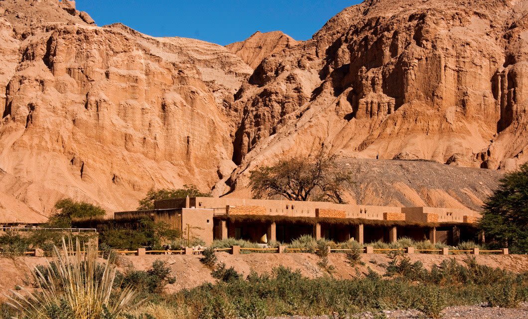 A modern building with earth-toned walls blends into the dramatic rocky cliffs and sparse desert vegetation of the Atacama Desert under a clear blue sky at Alto Atacama, Chile.
