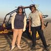 Two people wearing helmets stand smiling in front of a dune buggy on sandy terrain, ready for adventure and having the time of our lives under a clear blue sky.