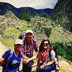 Three people wearing backpacks and outdoor clothing pose and smile on a hiking trail with ancient stone ruins and green mountains in the background, likely at Machu Picchu in Peru.