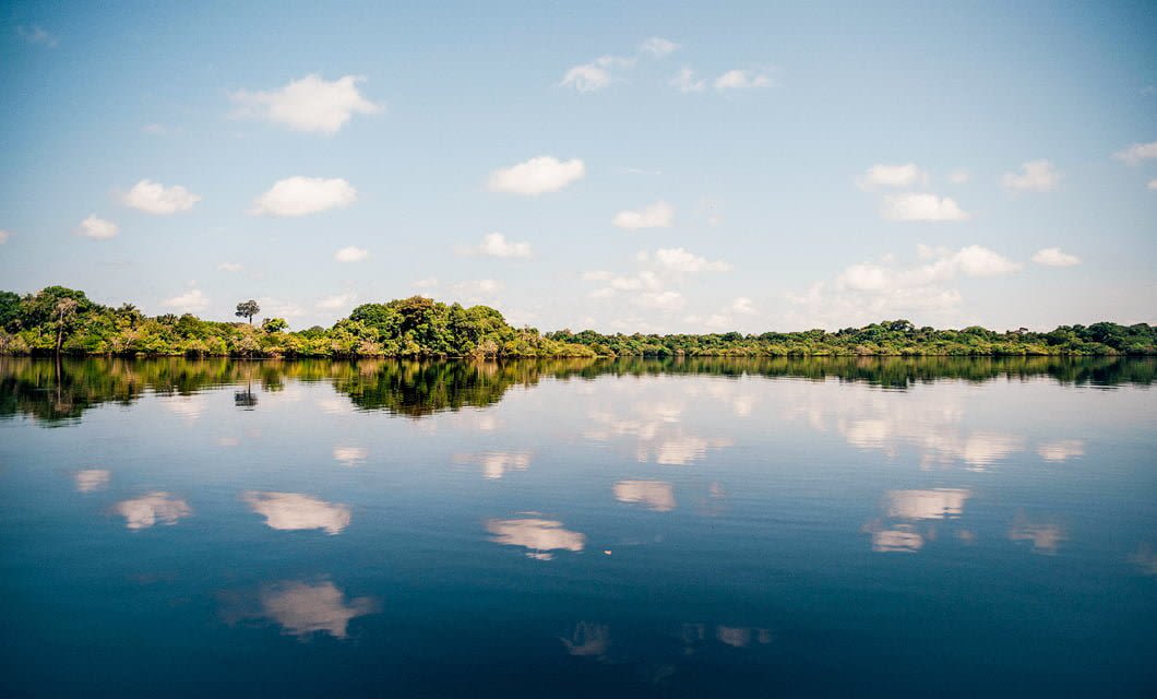 A calm river reflects a blue sky scattered with white clouds and the green trees lining the opposite shore, creating a serene and symmetrical landscape near Juma Amazon Lodge in the heart of the Amazon rainforest.