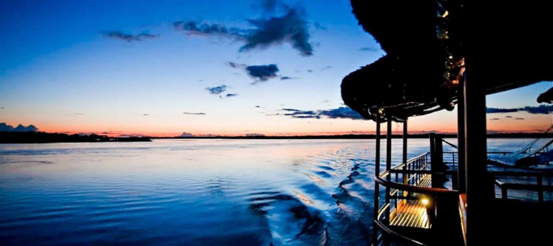 A boat moves across the wide Amazon River at sunset, leaving ripples in the water. The sky is a gradient of blue and orange, with scattered clouds and the boat’s lights reflecting on the surface—a scene from unforgettable Amazon River cruises.