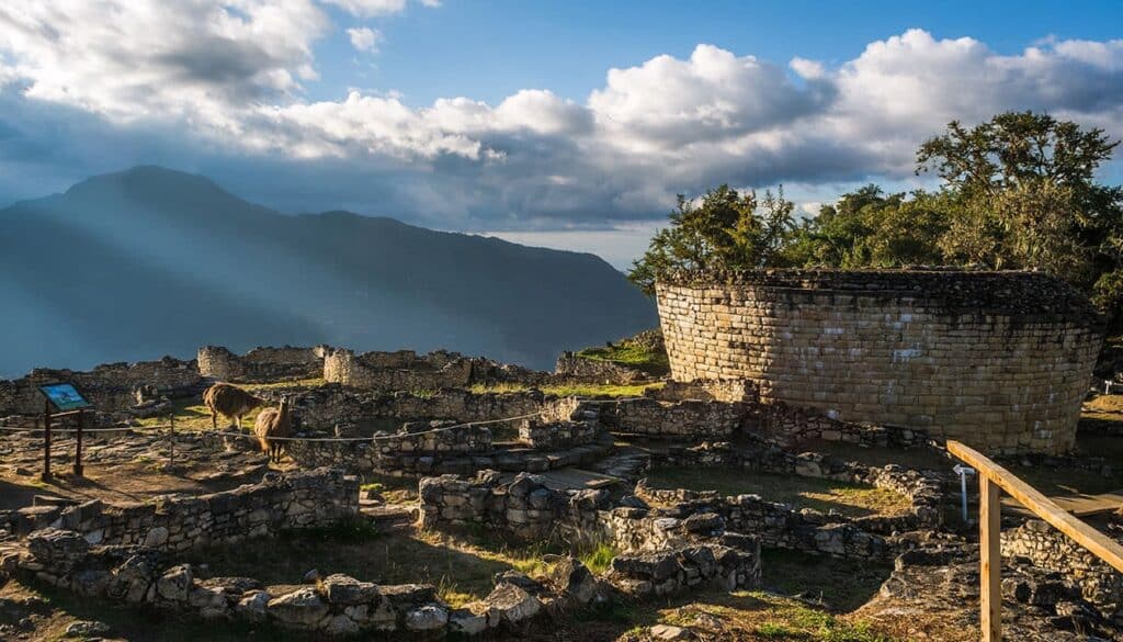 Ancient stone ruins on a mountain ridge in Peru, with a round tower, trees, and a dramatic cloudy sky. Sun rays illuminate the UNESCO landscape as distant mountains rise beyond—a llama grazes peacefully near the ruins.