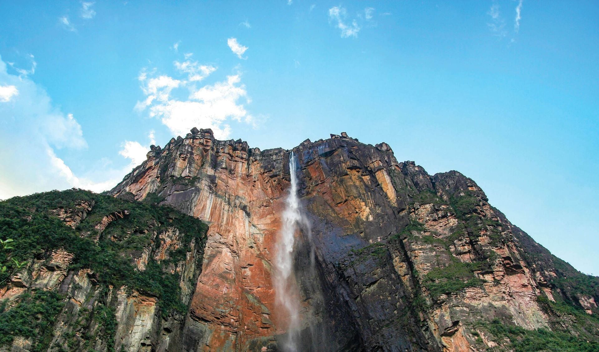 A tall waterfall cascades down a steep, rocky cliff covered with patches of greenery under a clear blue sky—an unforgettable sight for those exploring the Best Time to Visit Venezuela.