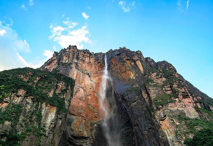 A tall, narrow waterfall cascades down a steep, rocky cliff covered in patches of green vegetation under a bright blue sky—one of the breathtaking Venezuela attractions perfect for your Venezuela travel adventure.