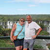 A middle-aged couple stands close together, smiling as they enjoy an enjoyable time in front of large waterfalls and lush greenery beneath a blue sky. The woman wears sunglasses and a turquoise top; the man sports a white shirt and shorts.