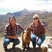 Two women kneel on a mountain path beside a brown alpaca, soaking in scenic mountains and a bright sky—capturing an authentic Peruvian experience.