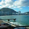 View of a city beach with a pier extending into the water, high-rise buildings along the shore, and a mountainous backdrop—an excellent job blending urban life with natural beauty under partly cloudy skies.