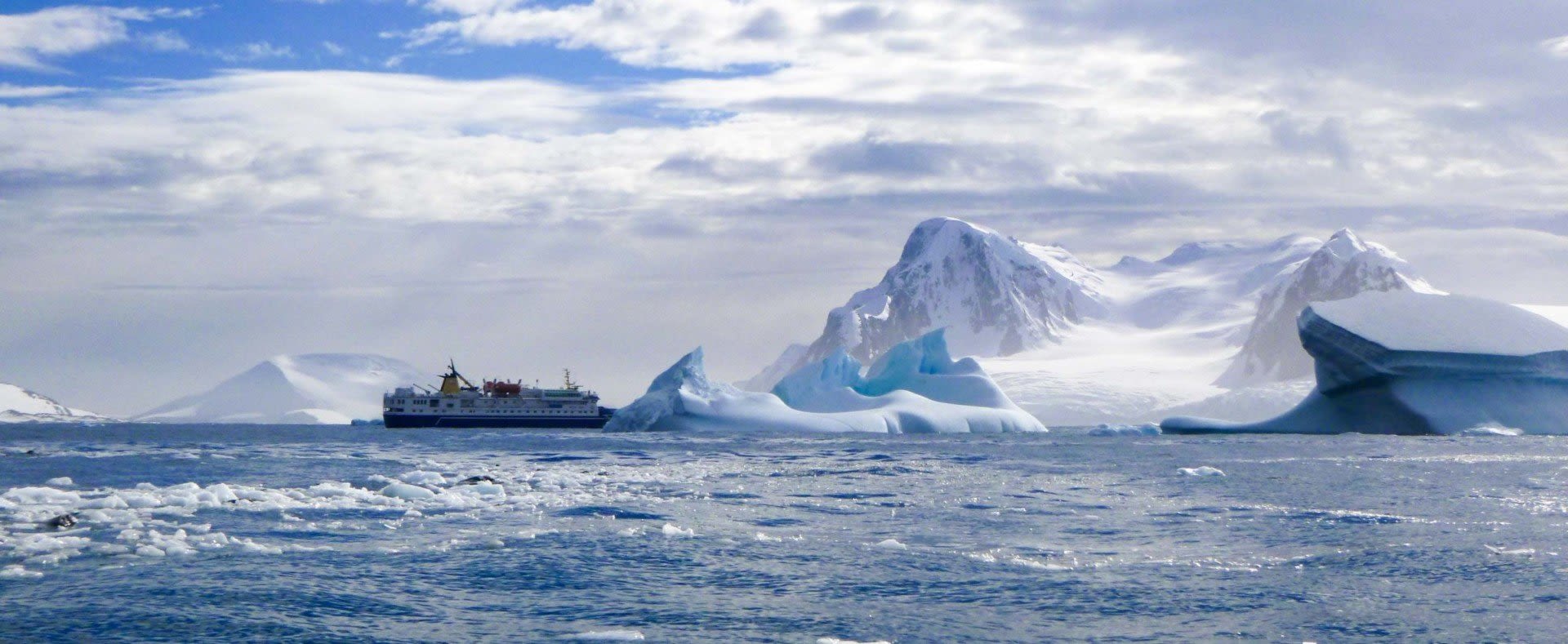 A ship sails through icy waters surrounded by floating icebergs, with snow-covered mountains and a cloudy sky in the background—an awe-inspiring scene that awaits those who embark on Antarctica travel after meeting all Antarctica visa requirements.