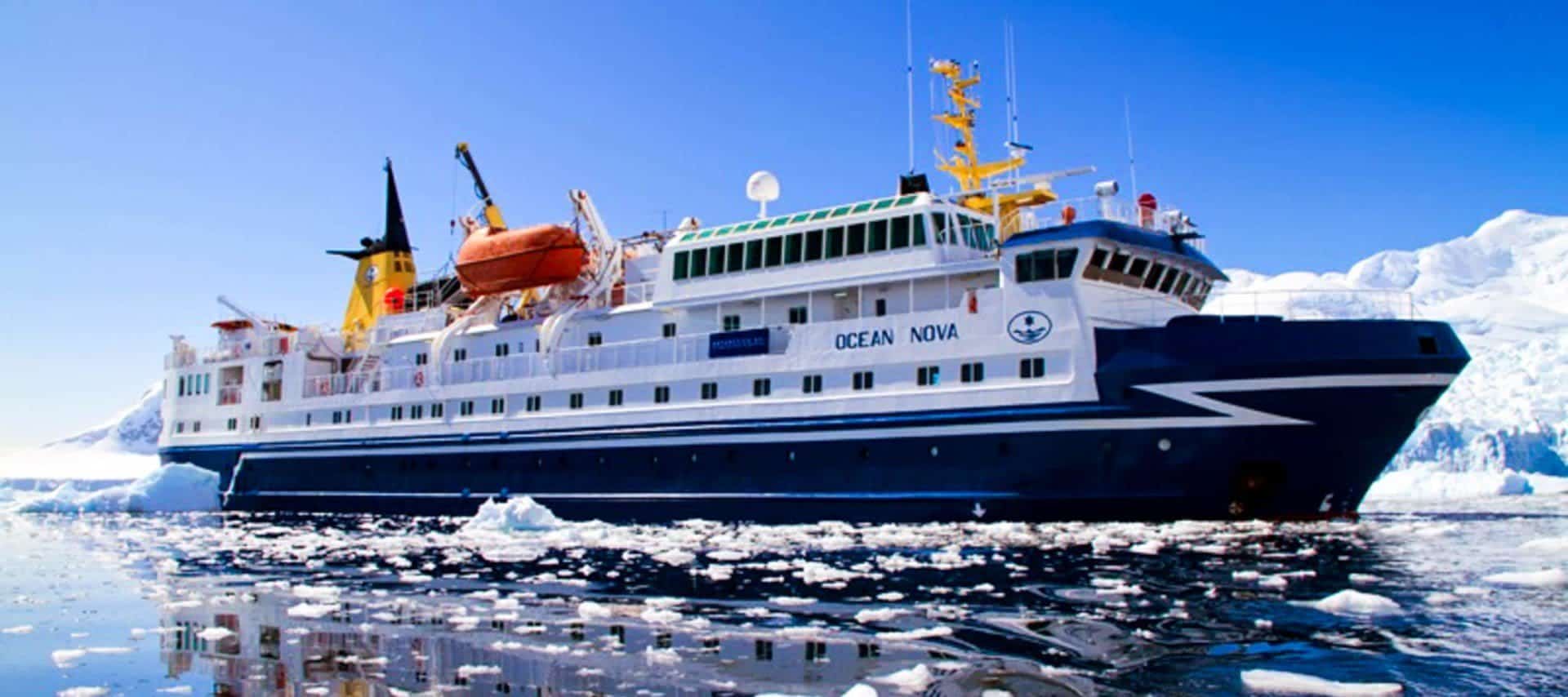 A large blue and white expedition ship named Ocean Nova sails through icy waters on one of the remarkable Antarctica Cruises, surrounded by floating ice and snow-covered landscapes under a clear blue sky.