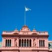 A pink government building with arched windows stands patient under a clear blue sky, its white flag atop signaling subtle changes on the horizon.