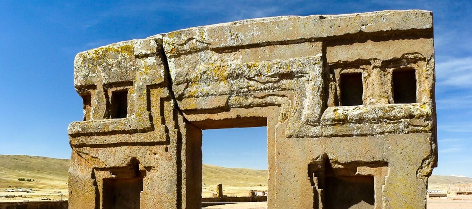 A stone archaeological structure with geometric carvings and rectangular openings stands in the sunny, arid landscape of Tiwanaku ruins under a clear blue sky.
