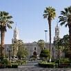 A historic stone cathedral with twin white towers stands behind a plaza lined with palm trees and trimmed bushes, promising a fine trip under the clear blue sky overhead.
