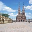 A large cobblestone plaza with a row of arches on the left and a tall twin-spired brick church in the background, under a blue sky with scattered clouds. People enjoy a fantastic time as they walk and gather in this lively South America setting.