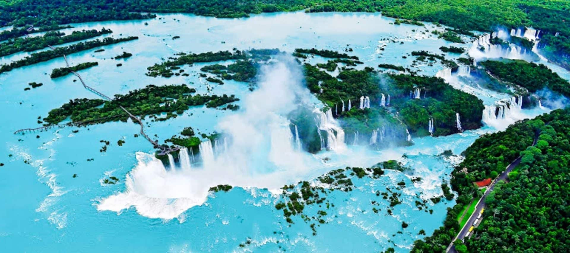 Aerial view of Iguazu Falls on the Brazil-Argentina border, with numerous waterfalls cascading into turquoise water, surrounded by lush rainforest and small islands—a breathtaking sight perfect for unforgettable tours under a bright sky.