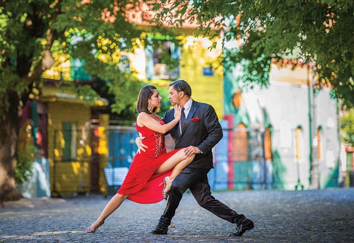 A couple dances tango outdoors on a cobblestone street—one of the classic things to do in Argentina. The woman wears a red dress and heels, the man a black suit. Colorful buildings and green trees are visible in the background.