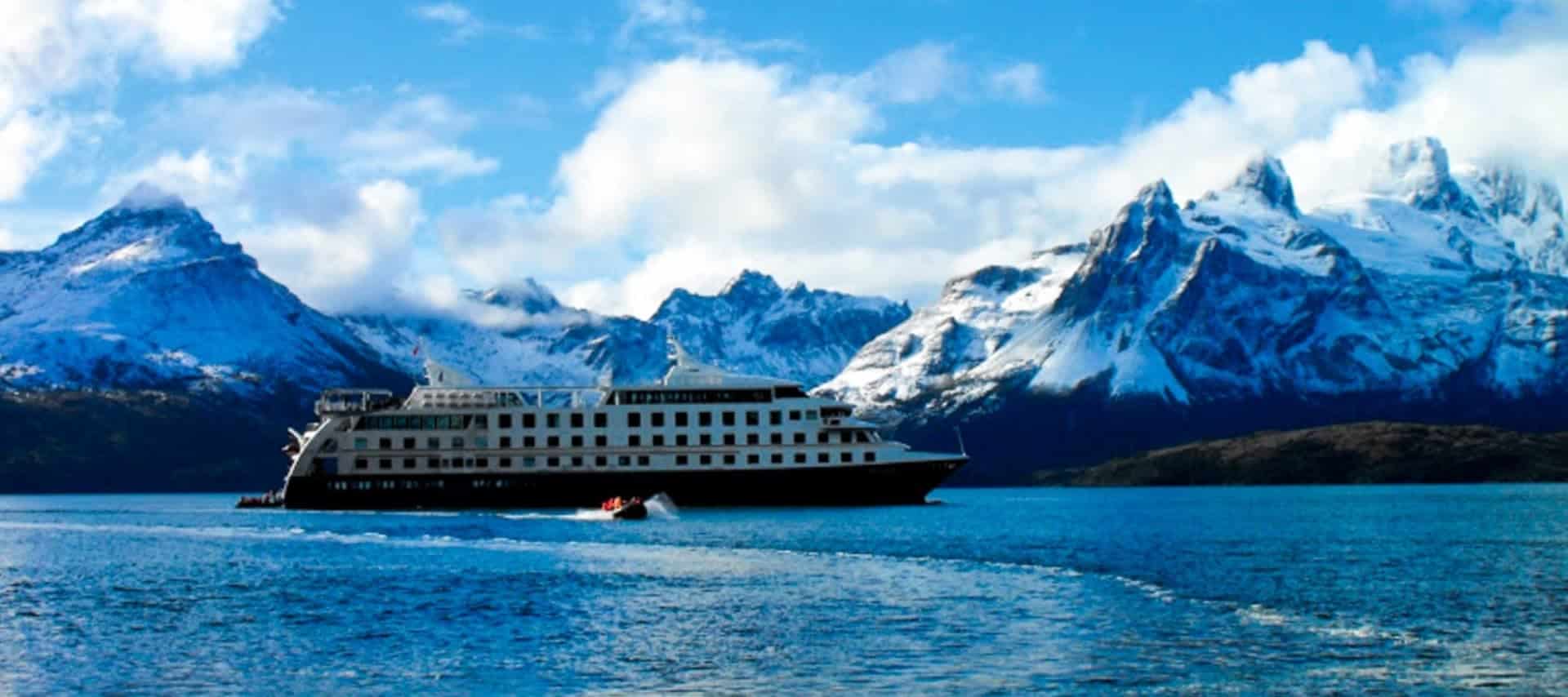 A large white cruise ship, part of Argentina Cruises, sails on a deep blue lake surrounded by snow-capped mountains under a partly cloudy sky. A small boat trails behind, leaving a wake in the water.