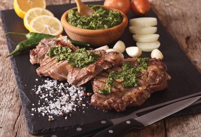 Three grilled steaks topped with green chimichurri sauce are served on a black slate board—a true taste of Argentina travel and one of the must-try Argentina attractions for food lovers. Garlic, lemon, onions, and peppers complete this authentic feast.