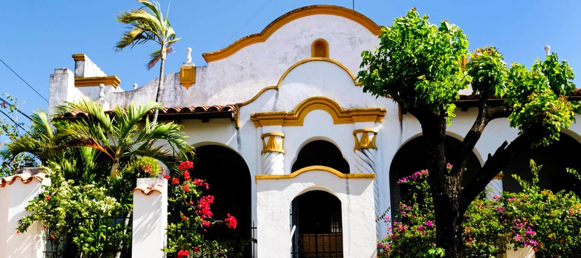 A vibrant, white stucco building with yellow trim, arched doorways, and a tiled roof—typical of Jesuit Missions—stands amid lush green trees and flowering plants under a bright blue sky. Perfect for those seeking Jesuit Missions Tours to Paraguay.