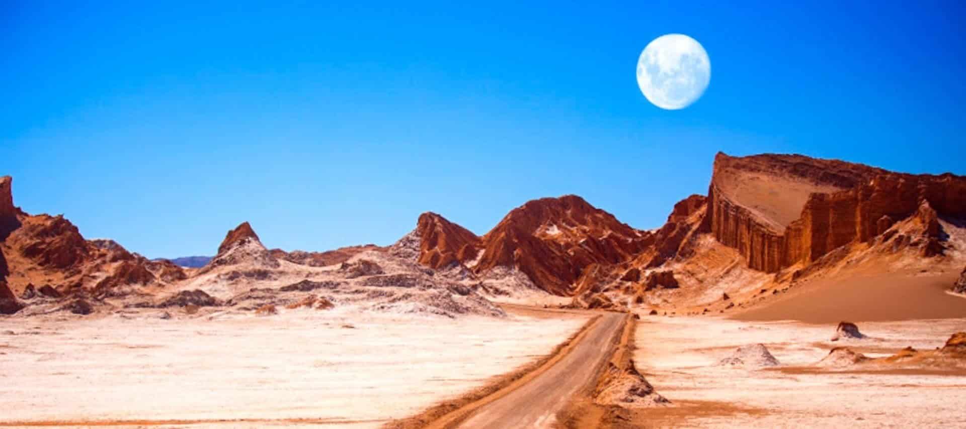 A dirt road winds through the dry, rocky Atacama Desert with reddish-brown hills under a bright blue sky. A large, white full moon rises above the rugged landscape, perfect for unforgettable Atacama Desert tours.
