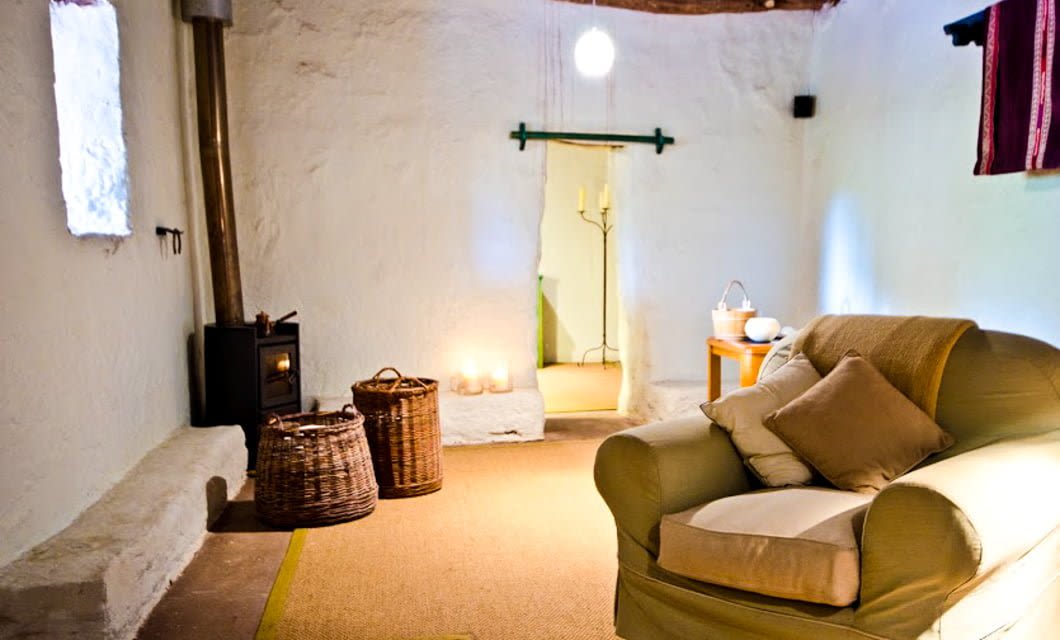 A cozy room with white plaster walls, a cushioned armchair with pillows, a wood-burning stove, wicker baskets, candles, and a small table—echoing the serene comfort of Explora Atacama in the heart of the Atacama Desert.
