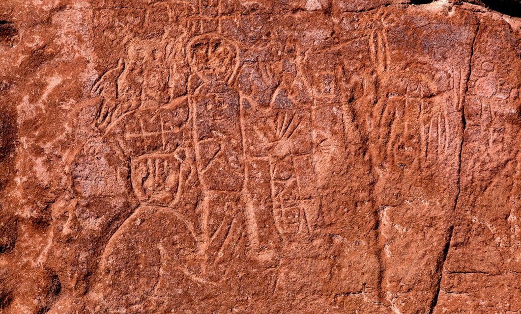 Ancient petroglyphs carved into a reddish-brown rock surface in the Atacama Desert, depicting human-like figures and abstract shapes, with weathered textures and natural cracks visible near Alto Atacama.
